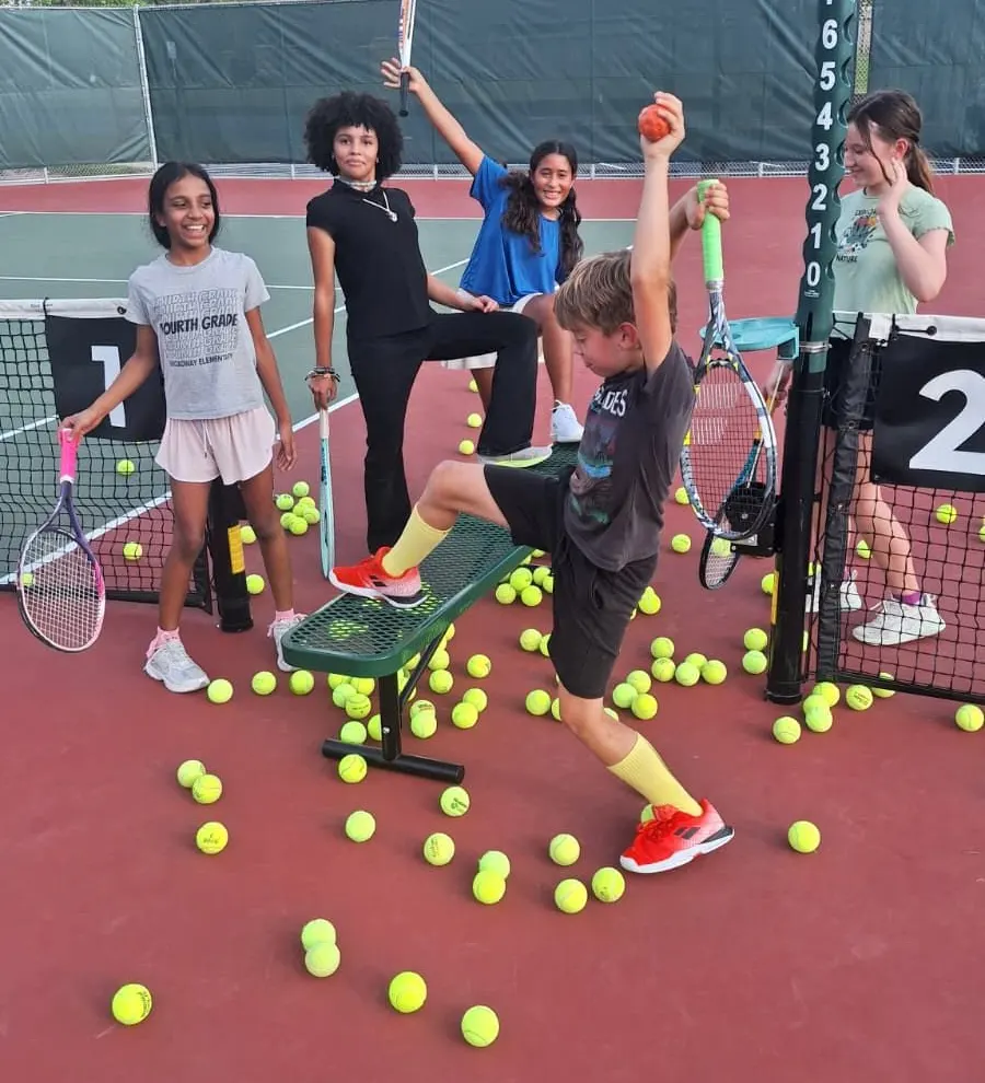Young players laughing as one kicks through a pile of tennis balls spread across the court.