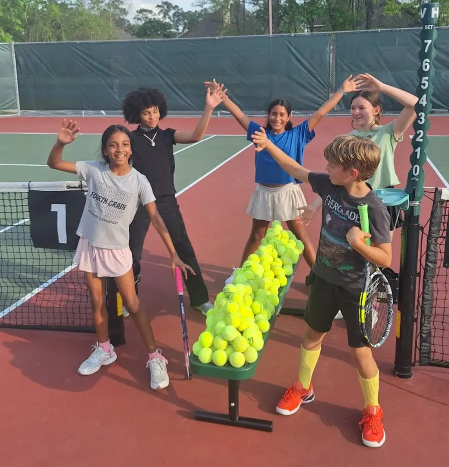 A group of young players raising their hands beside a tall stack of tennis balls at the net.