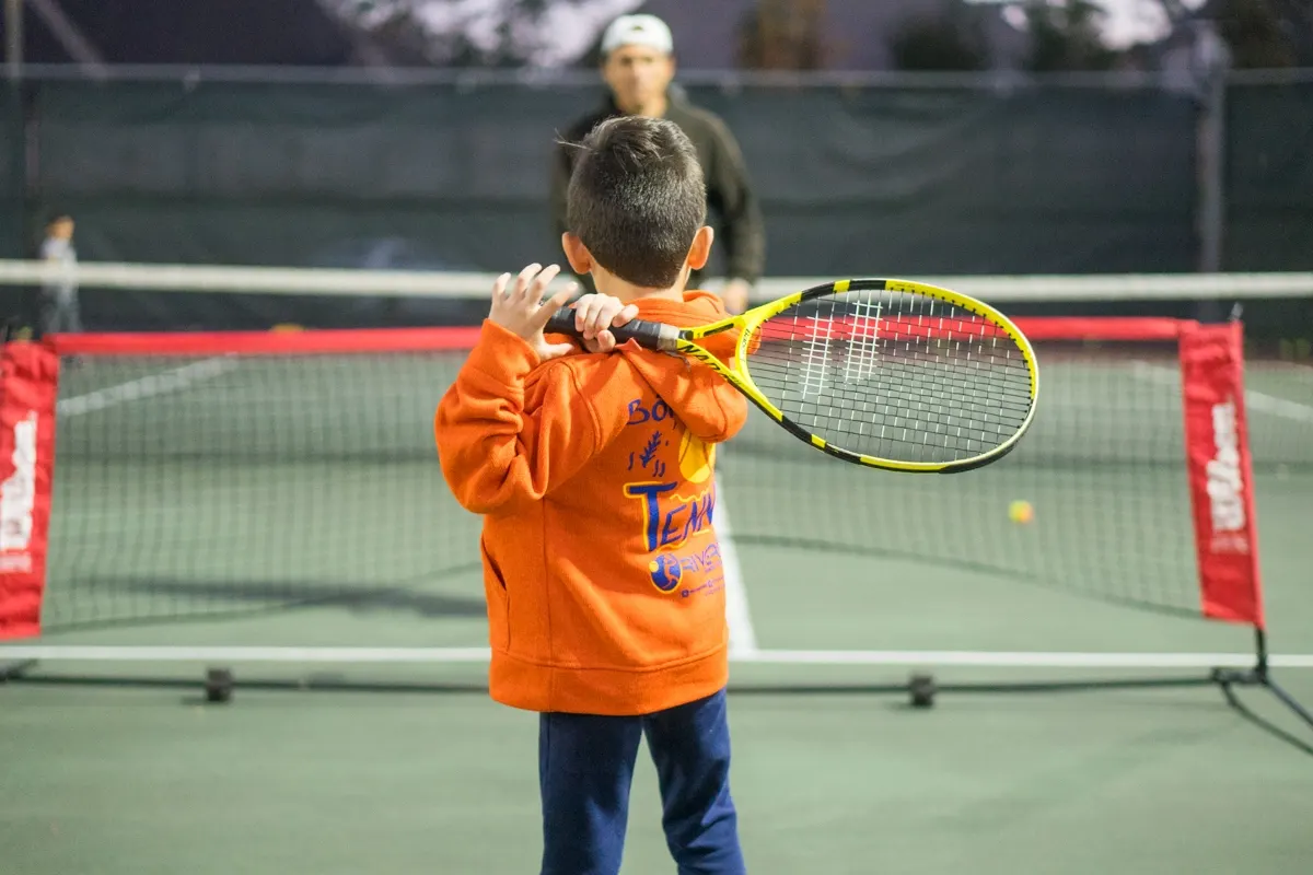 A young child in an orange Rivera Tennis Academy hoodie standing at a red mini net, racquet resting on one shoulder, a coach waiting on the far side.