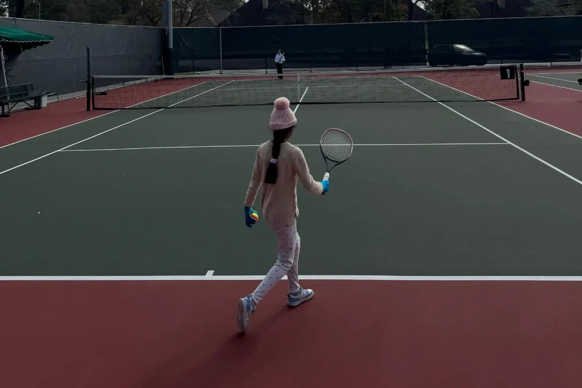 A child in a knit hat walking up the court with a racquet and an orange low-compression ball, portable net behind.