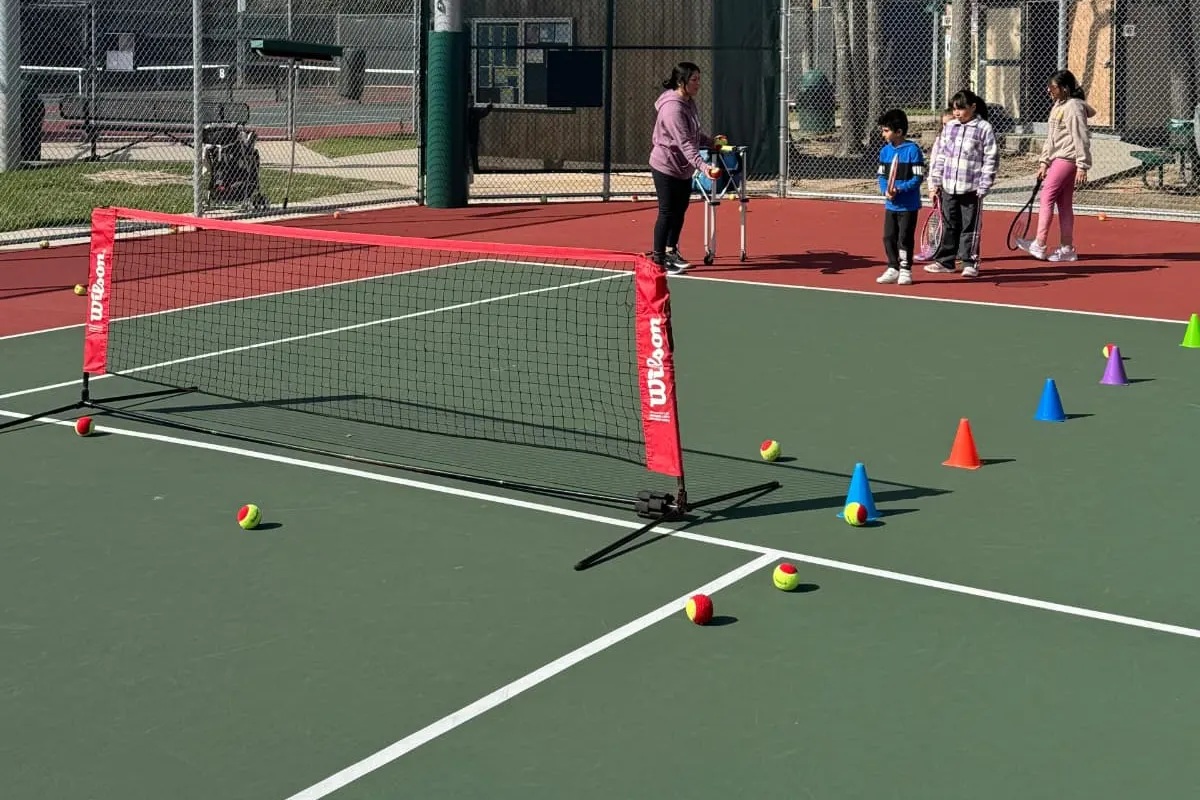 A red mini net set up across the court with red low-compression balls and coloured cones, a coach at the ball cart and three children waiting with racquets.