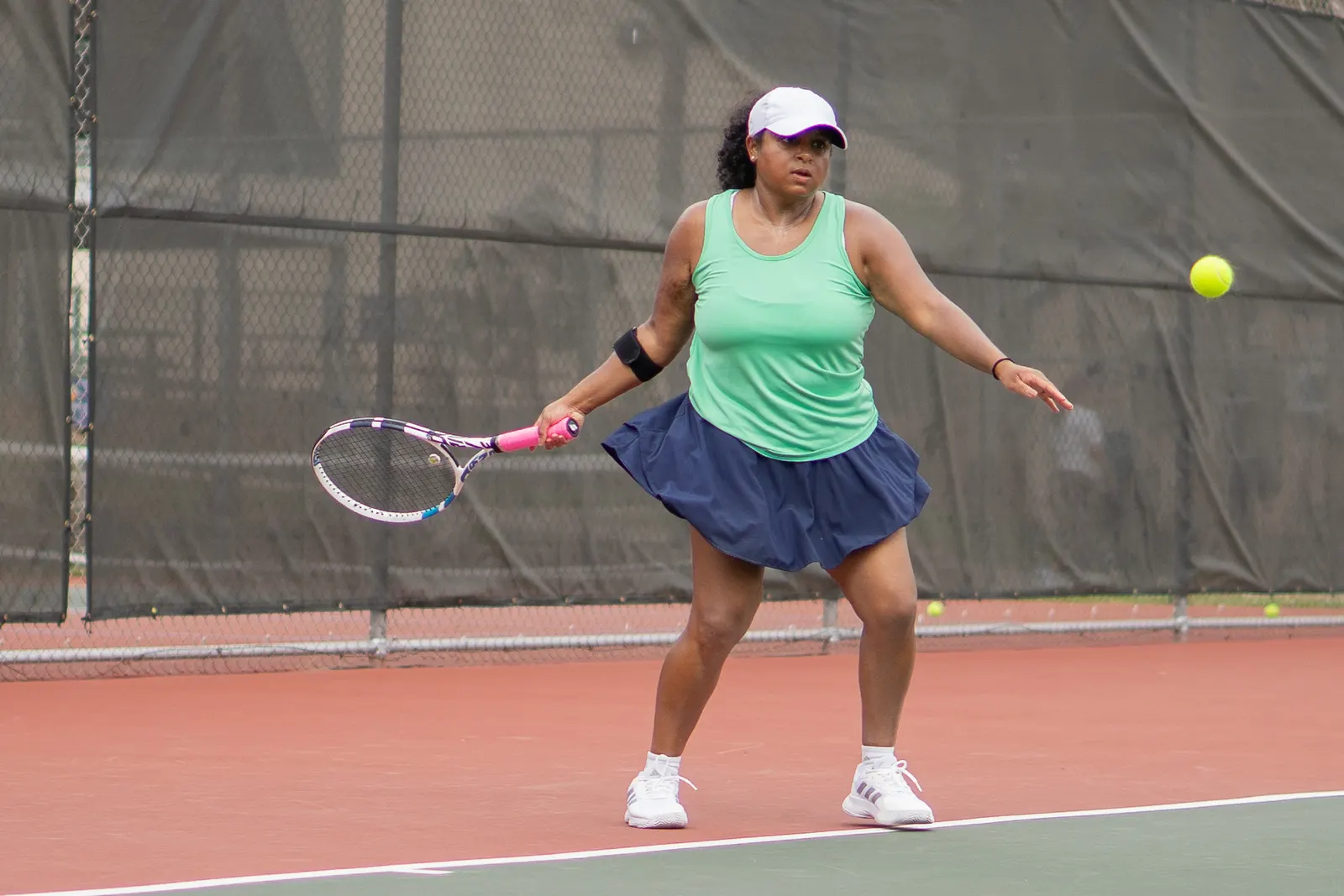 Player in a green tank top and white cap on a hard court, racquet drawn back low to her forehand side.
