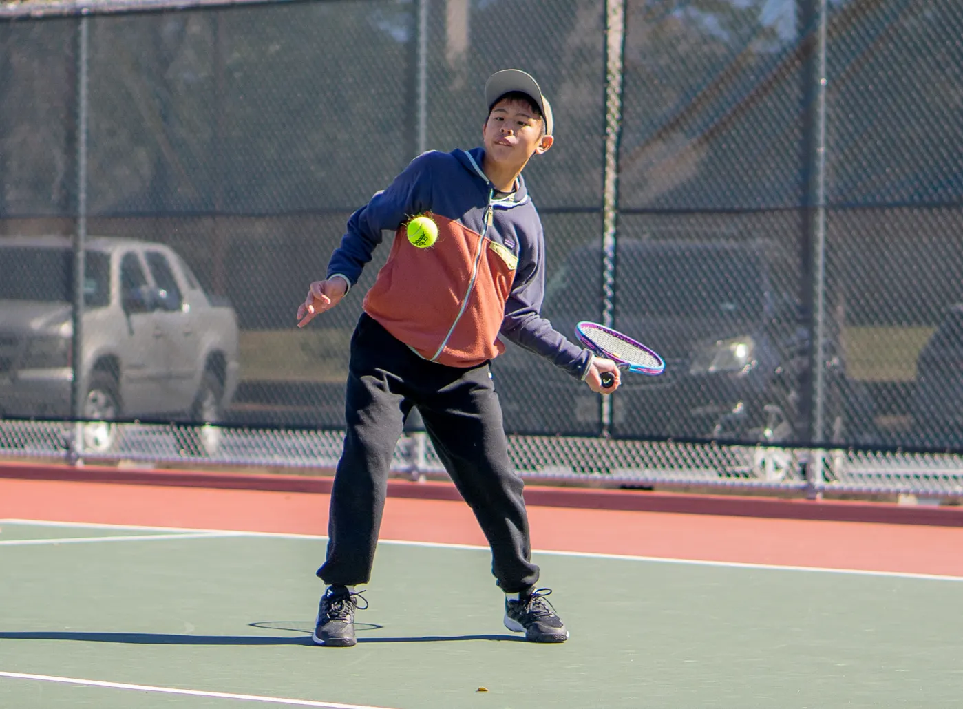 Junior player in a cap and hoodie on a hard court with racquet drawn back low and a ball in the air.
