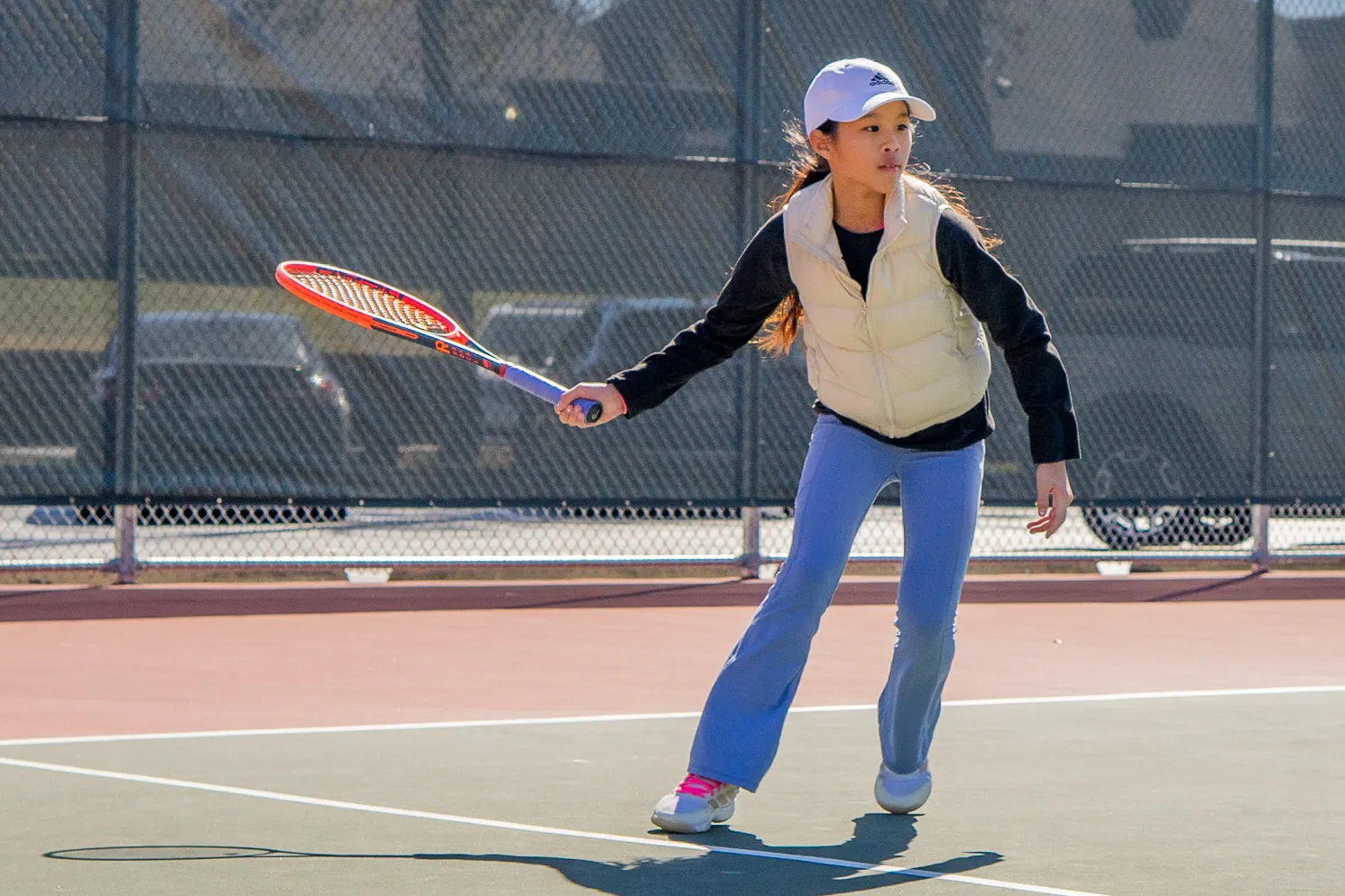 Junior player in a white cap and puffer vest moving forward with the racquet extended on an outdoor court.