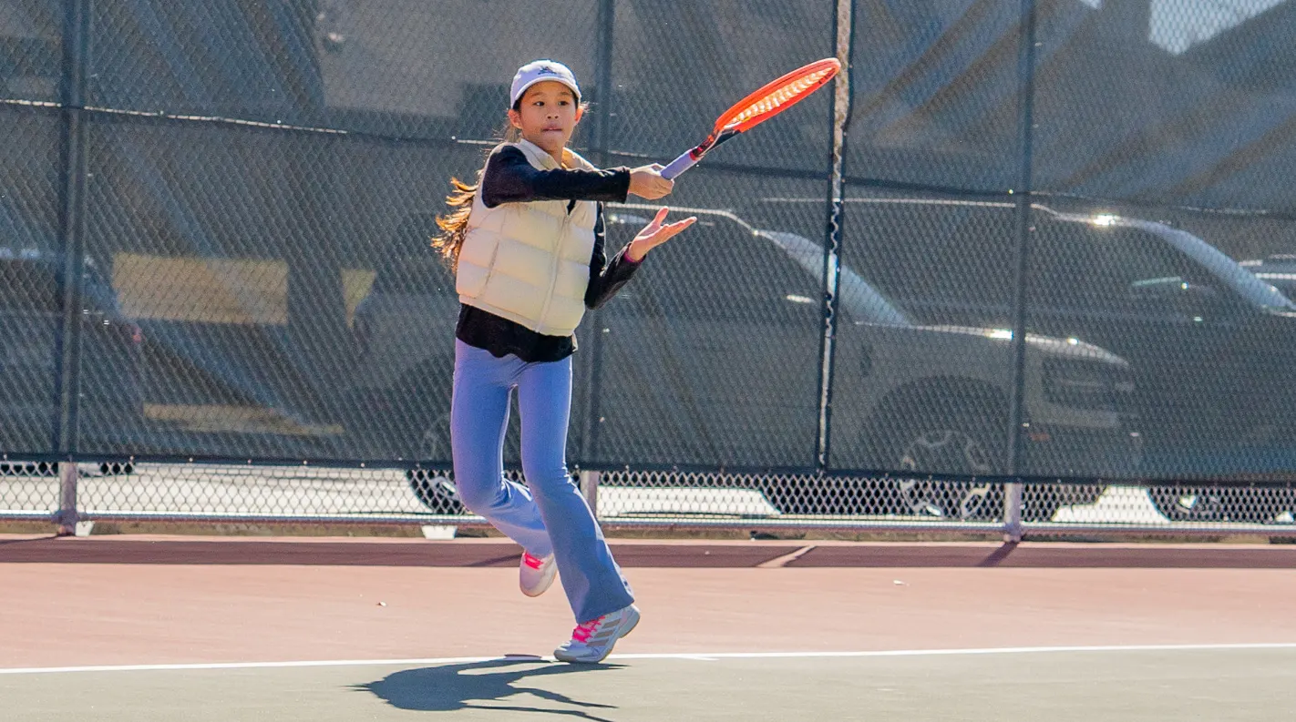 Young player leaping with racquet raised overhead as a tennis ball flies high above a hard court.