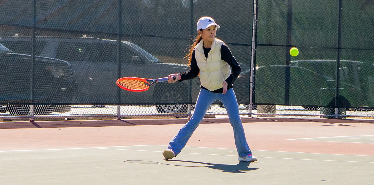 Young player reaching low with the racquet to one side on a hard court, ball in the air nearby.