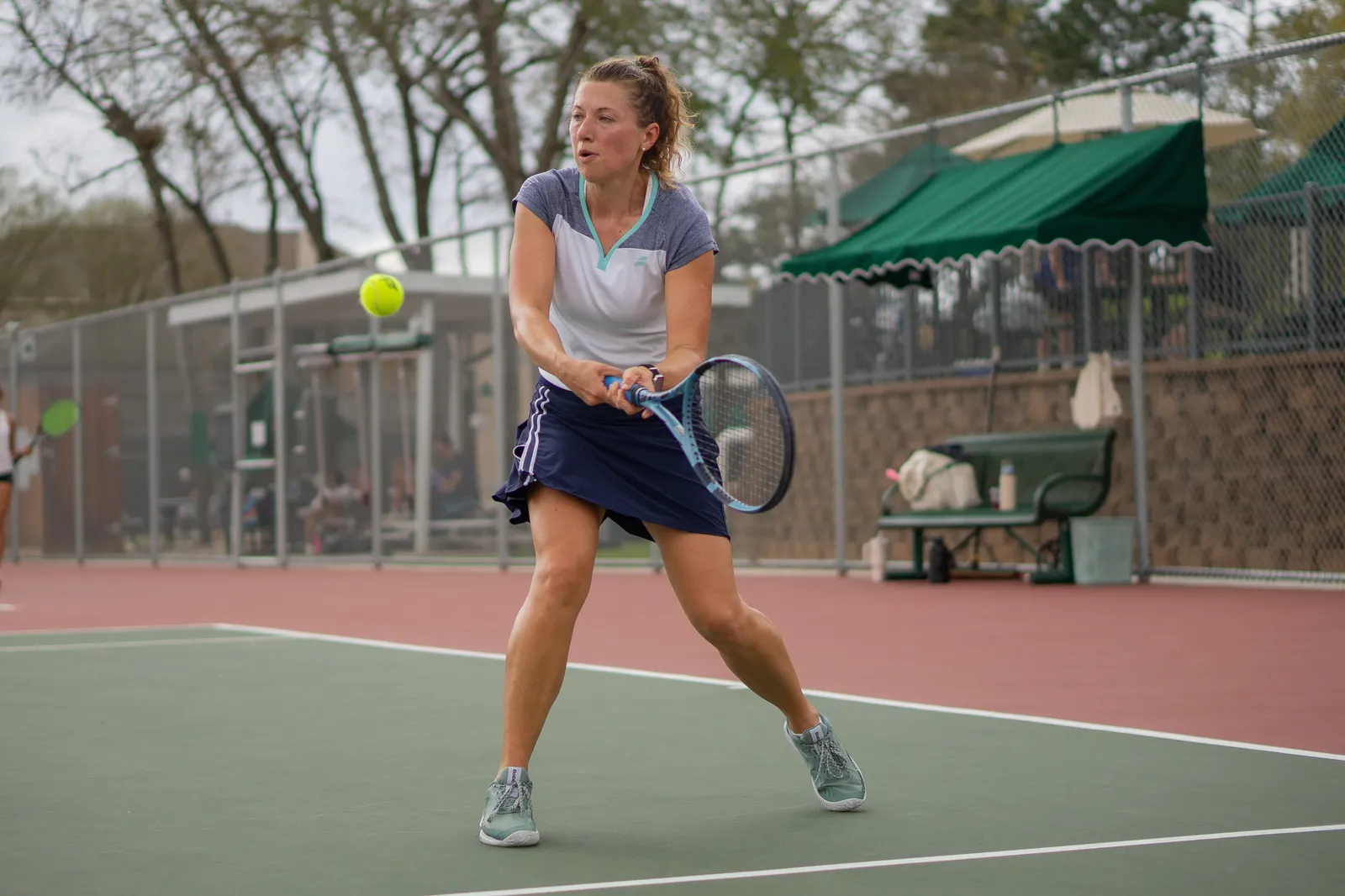 Player taking the racquet back with both hands for a backhand on an outdoor hard court, ball in the air.