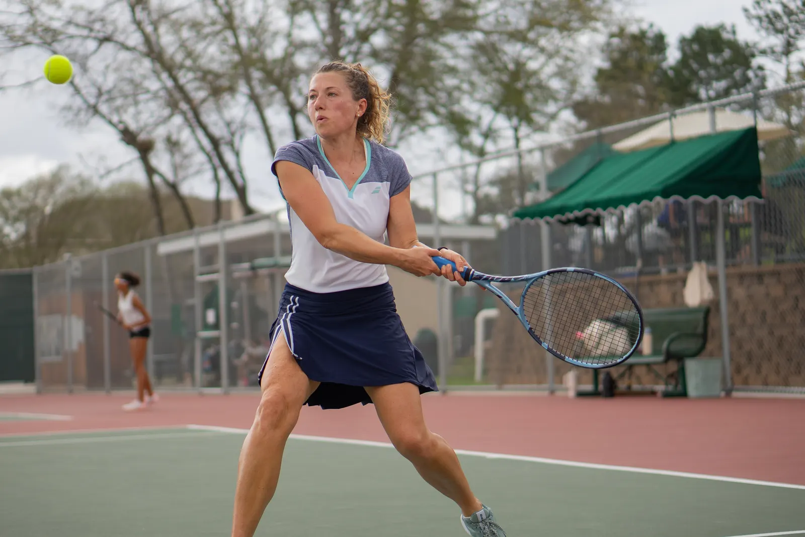 Player hitting a two-handed backhand on an outdoor hard court, arms extended through the swing.