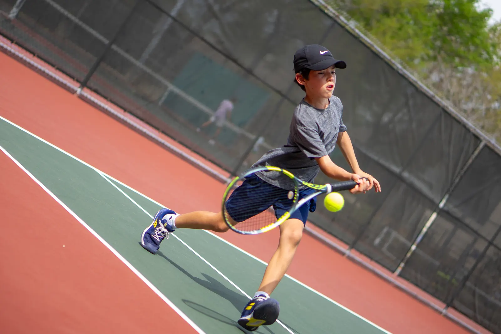 Young player stretched low for a one-handed backhand, ball at the strings, on an outdoor hard court.