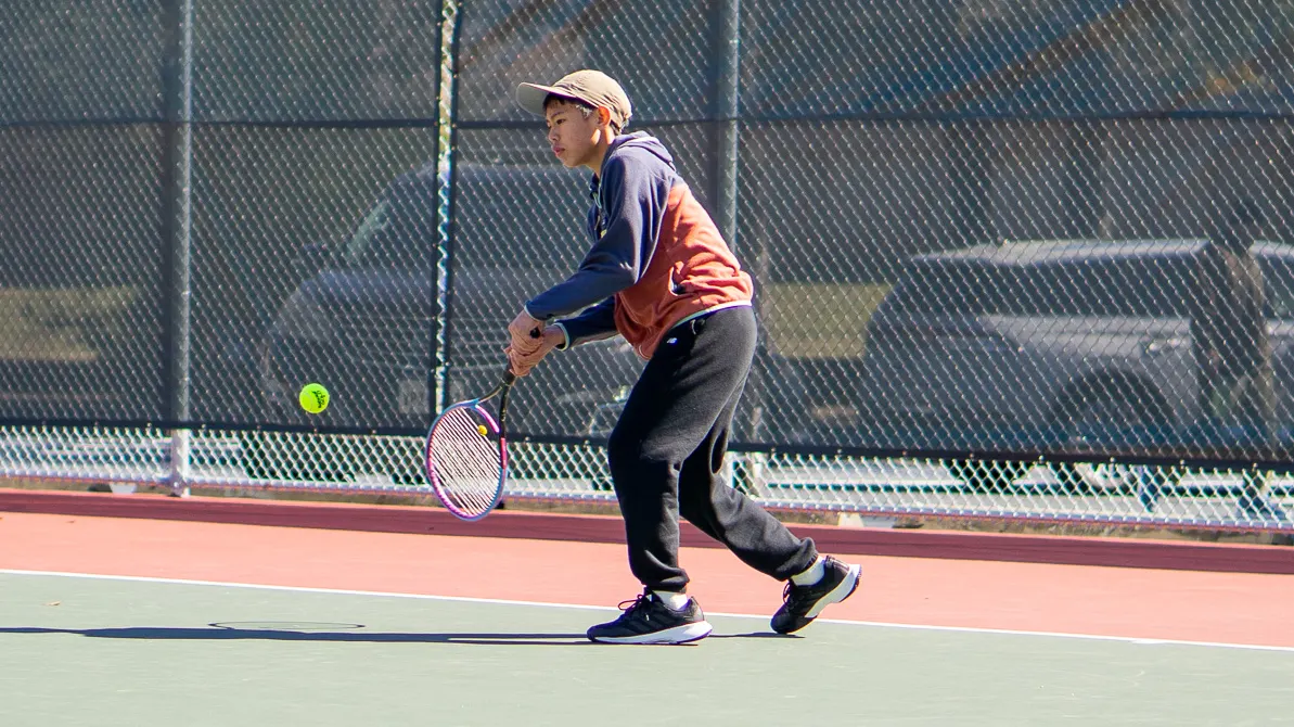 Junior player in a tan cap hitting a low two-handed backhand on an outdoor hard court.