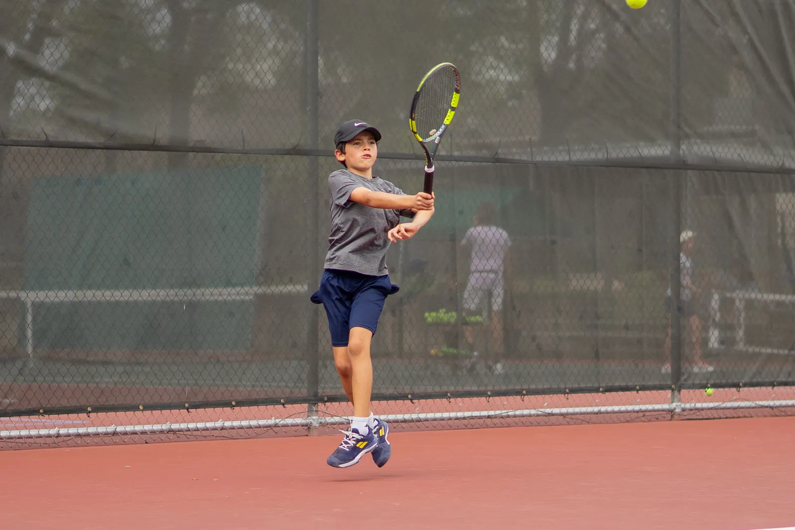 Young player leaving the ground on a hard court, racquet finishing high, ball in the air.
