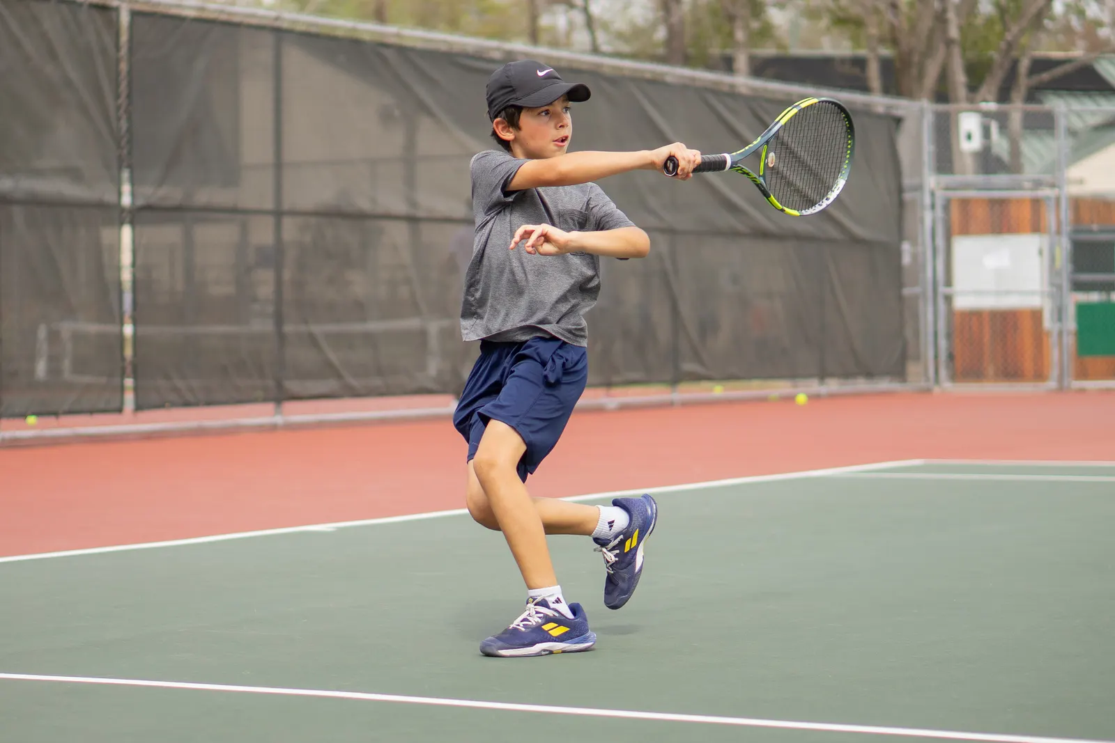 Young player in a cap hitting a forehand on an outdoor hard court, airborne with feet off the ground