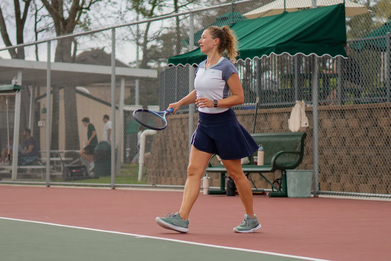 Player walking across an outdoor hard court holding a racquet, between points.