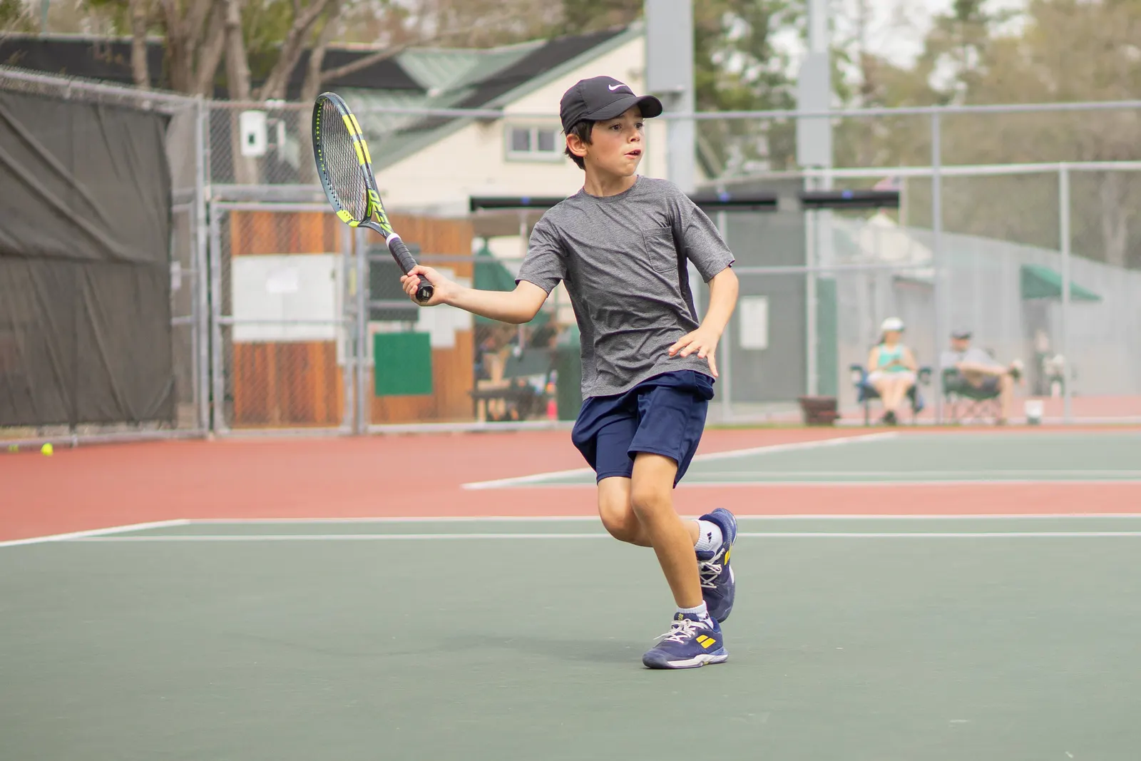A child running across an outdoor hard court with racquet in hand as a ball flies to one side.