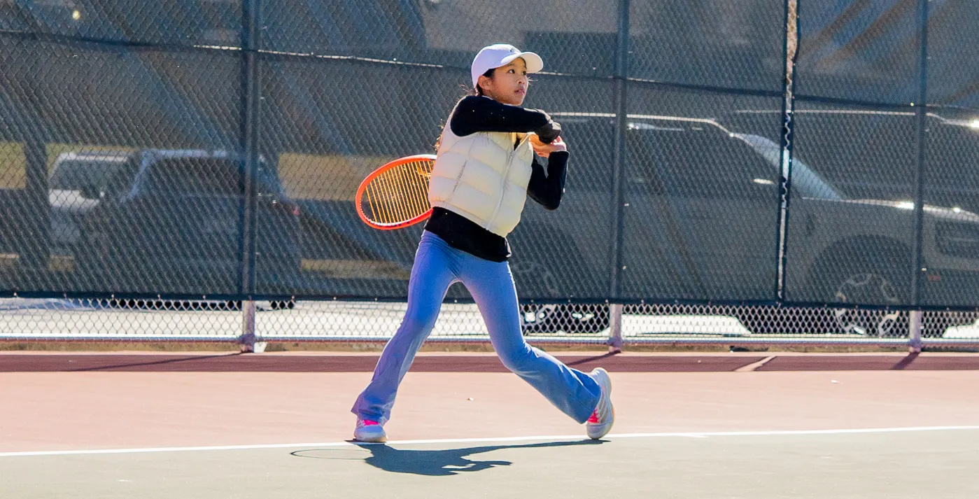 Young player in a cap striding across a hard court gripping the racquet with both hands.
