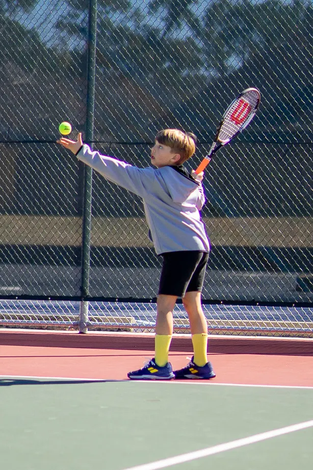Young child tossing the ball to serve, racquet raised behind, standing at the baseline.